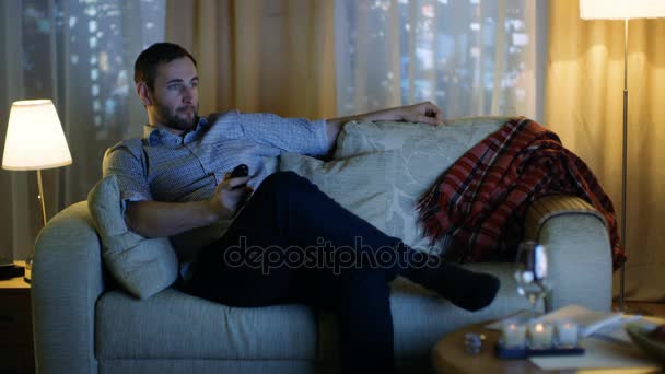 夜中は、テレビを見てソファの上に座っている老人は、リモコンでチャンネルを変更します。大都市では高層ビルの背景ウィンドウ ビューで. — ストック動画