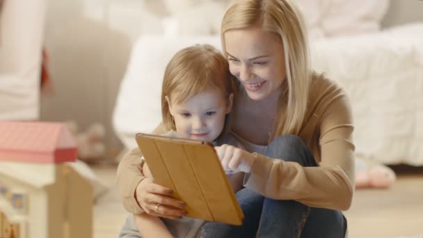 Krásná matka a její malá dcera mají dobrý čas čtení dětských knih v tabletovém počítači. Dětský pokoj je plný hraček. — Stock video