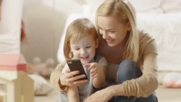 Mooie jonge moeder met haar dochtertje zit en toont haar iets interessants op een Smartphone. Kinderkamer zit vol met speelgoed. — Stockvideo