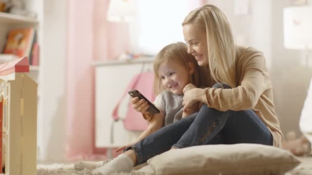 Hermosa madre joven se sienta con su hijita y le muestra algo interesante en un teléfono inteligente. Habitación de los niños es de color rosa y lleno de juguetes . — Vídeos de Stock