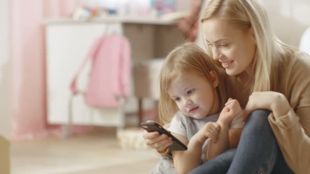 美しい若い母親と彼女の小さな娘座ってスマート フォンで彼女の何か面白いものを示しています。子供の部屋はピンクとフルのおもちゃ. — ストック動画