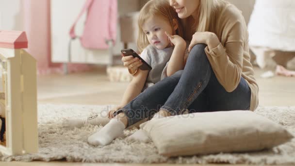 Bella giovane madre si siede con la sua piccola figlia e le mostra qualcosa di interessante su uno smartphone. La stanza dei bambini è rosa e piena di giocattoli . — Video Stock