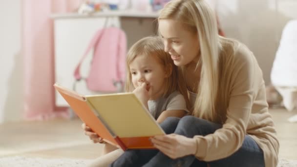 Belle jeune mère et sa jolie petite fille lisent ensemble le livre pour enfants. La chambre des enfants est rose et pleine de jouets . — Video