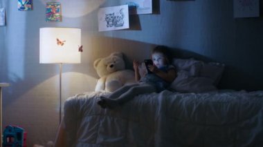 Sevimli küçük kız yalanlar yatak ve saat karikatürler bir smartphone. Onun zemin lamba yanıyor.