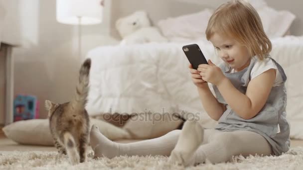 Nettes kleines Mädchen sitzt mit Smartphone auf dem Boden und filmt ihr gestreiftes Kätzchen beim Herumlaufen. Zeitlupe. — Stockvideo
