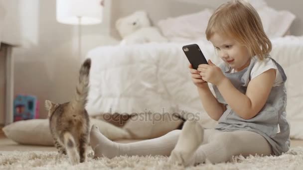 かわいい女の子は、スマート フォンや彼女の縞模様の子猫の歩き回る撮影ビデオで床に座っています。スローモーション. — ストック動画