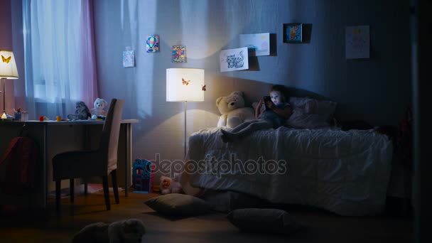 Petite fille mignonne repose sur son lit et regarde des dessins animés sur un smartphone. Son chaton est à côté de son jeu. Sa lampe de sol est allumée . — Video