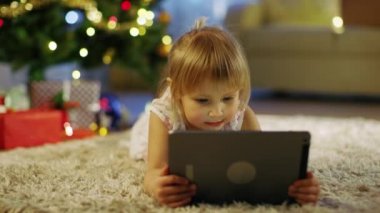 Sevimli küçük kız ellerini Tablet bilgisayar ile Noel ağacı altında halı üzerinde yatıyor.