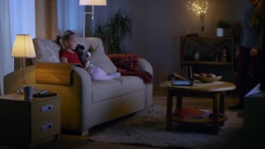 Akşam küçük kız bir oturma odasında bir kanepede oturuyor. O onun yumuşak oyuncak ve saatler Tv, onu güzel anne birleşimler geldi ve onu bir kanepe ve katıldı tutar.