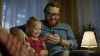 Akşam baba ve küçük kız oturan bir kanepe oturma odası seyretmek smartphone cep telefonu ile komik videolar.