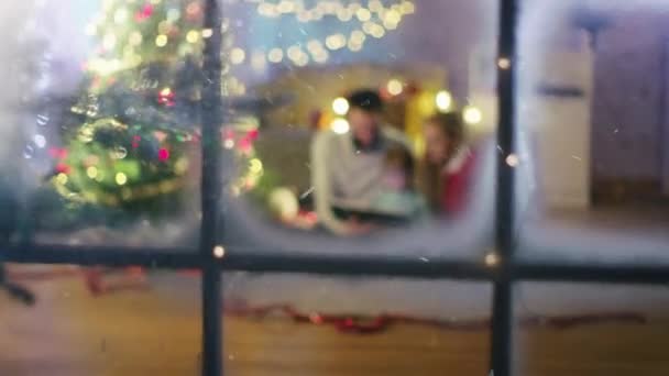 Κοιτάζοντας μέσα από το παράθυρο. Κάθεται κάτω από το χριστουγεννιάτικο τρία δώρα κάτω από το Πατρός και μητρός Διαβάστε ένα βιβλίο για τη μικρή της κόρη. — Αρχείο Βίντεο