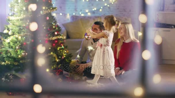 Regardant par la fenêtre enneigée. Famille heureuse : Père, Mère et leur petite fille décorent l'arbre de Noël . — Video