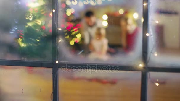 Κοιτάζοντας μέσα από το χιονισμένο παράθυρο. Ευτυχής πατέρας, μητέρα και κόρη κάθεται κάτω από το χριστουγεννιάτικο δέντρο. Κόρη ανοίγει τα δώρα. — Αρχείο Βίντεο