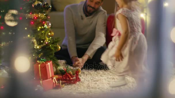 Belenéz a havas ablak. Boldog apa, anya és lánya ül a karácsonyfa alatt. Lánya ad egy ajándék az anyjával. — Stock videók