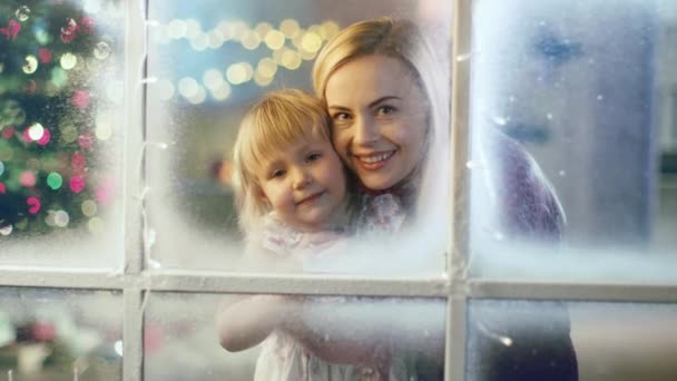 Την παραμονή των Χριστουγέννων μητέρα και κόρη, κοιτάζοντας μέσα από το χιονισμένο παράθυρο. Γιρλάντα λάμπει φωτεινό σε ένα παράθυρο. — Αρχείο Βίντεο