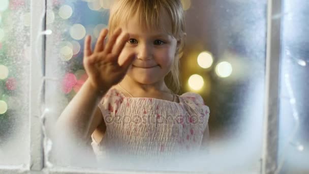 Na véspera de Natal bonito menina olha através do nevado, janela congelada e sorrisos . — Vídeo de Stock