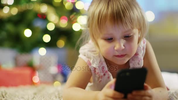 Милая маленькая девочка со смартфоном в руках лежит на ковре под елкой . — стоковое видео