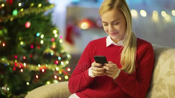 Hermosa mujer rubia se sienta en un sofá y utiliza Smartphone. Árbol de Navidad y habitación decorada con luces están en el fondo . — Vídeo de stock