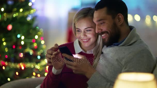 Homem bonito e mulher bonita sentado no sofá usando Smartphone. Homem segura Sua Senhora. No fundo, muitas luzes e árvores de Natal . — Vídeo de Stock