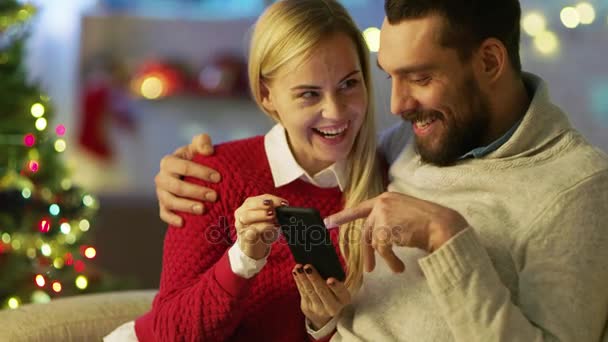 Pohledný muž a krásná žena sedí na gauči, pomocí Smartphone. Muž drží jeho dáma. V pozadí mnoha světel a vánoční stromeček. — Stock video