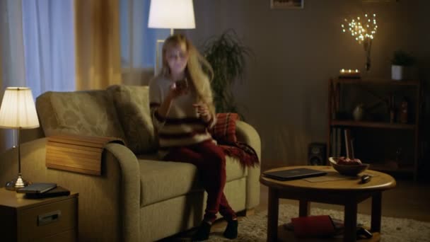Piękna młoda kobieta wchodzi pokój dzienny w godzinach wieczornych. Siedzi na jej kanapie i zaczyna się przy użyciu smartfona. — Wideo stockowe