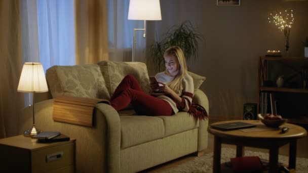 En la noche hermosa mujer joven se encuentra en el sofá y utiliza el teléfono inteligente. Habitación se ve cálido y acogedor . — Vídeos de Stock