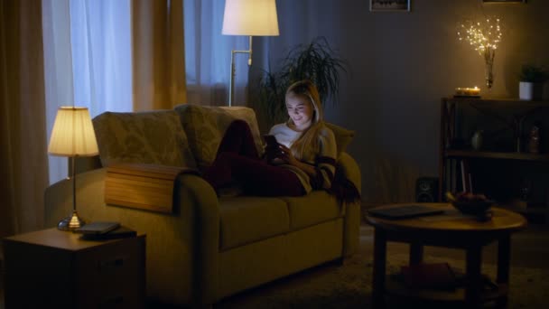 In the Evening Beautiful Young Woman Lies on the Couch and Uses Smartphone. Room Looks Warm and Cozy. — Stock Video