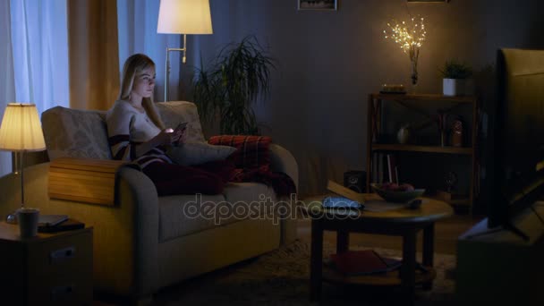 In the Evening Beautiful Young Woman Relaxes on a Couch in Her Cozy Living Room. She Uses Her Smartphone. — Stock Video