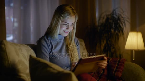 Vacker ung kvinna i hennes vardagsrum. Hon sittandes på en soffa och använder Tablet PC: N. Bakom hennes stora staden syns i fönstret. — Stockvideo