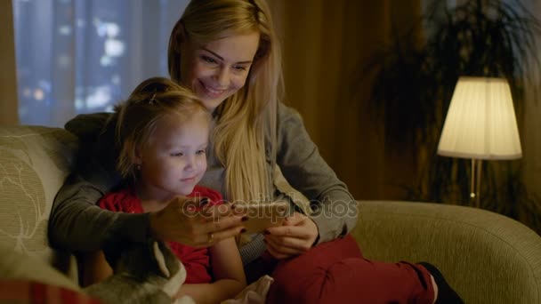 Anne ve kız şirin kızı oturan üzerinde bir kanepe oturma odasında vardır. Onlar kullanma Smartphone ve gülümseyerek. Akşam oldu. — Stok video