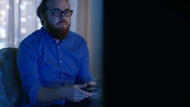 Akşam adam kanepede oturuyor ve video oyunları içinde onun oturma odasında oynuyor. Windows görüldü arkasında büyük şehirdir.