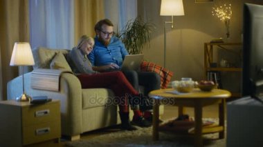 Kız ve erkek dizlerinin üstünde Laptop ile bir kanepede oturuyor, video izleme ve Having Fun. Rahat oturma odam. Akşam oldu.