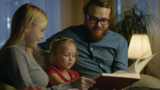 Padre, madre e hijita leyendo el libro infantil sobre un sofá en la sala de estar. Es de noche. . — Vídeo de stock