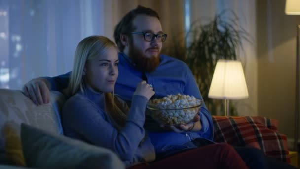 Pojkvän och flickvän tittar på Tv. De sitter på en soffa i sin mysiga vardagsrum och äta Popcorn. Det är kväll. — Stockvideo