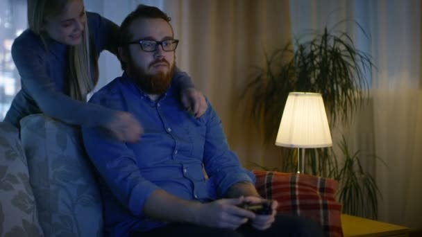 En el hombre de la tarde sentado en un sofá jugando videojuegos, su cónyuge entra y lo abraza . — Vídeo de stock