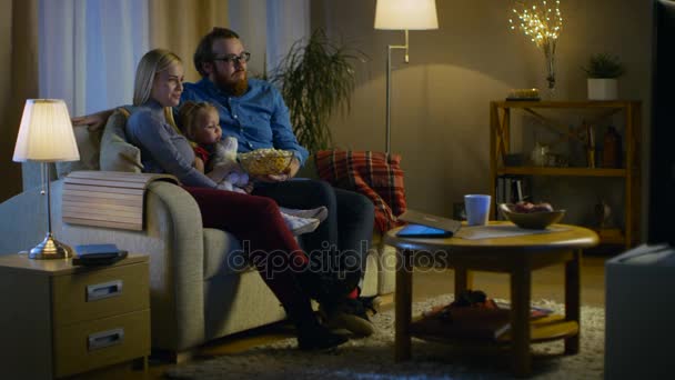 Long Shot of a Father, Mother and Little Girl Watching TV. Se sientan en un sofá en su acogedor salón y comen palomitas de maíz. Es de noche. . — Vídeo de stock