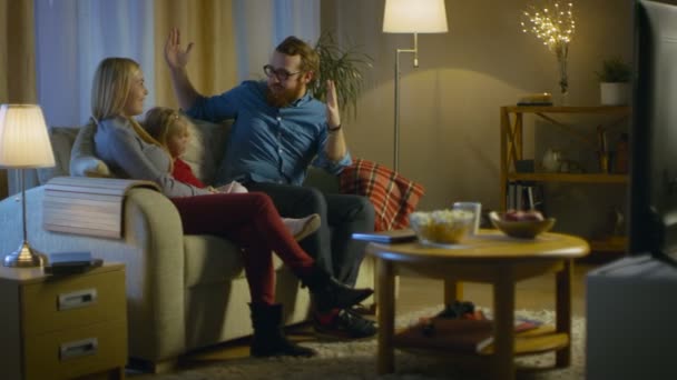 Lange Aufnahme eines Vaters, einer Mutter und eines kleinen Mädchens vor dem Fernseher. Vater erklärt seinem kleinen Mädchen etwas. Sie sitzen auf einem Sofa in ihrem gemütlichen Wohnzimmer. Es ist Abend. — Stockvideo