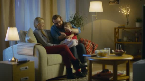 Long Shot of a Father, Mother and Little Girl Watching TV. Father Explains Something to His Little Girl. They Sit on a Sofa in Their Cozy Living Room. It's Evening. — Stock Video