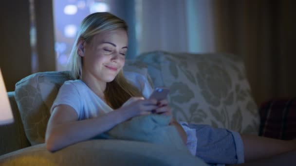 A tarda notte Bella donna sdraiata sul suo divano in salotto, usando il suo smartphone e sorridendo. Il suo appartamento ha grande vista sulla finestra della città . — Video Stock