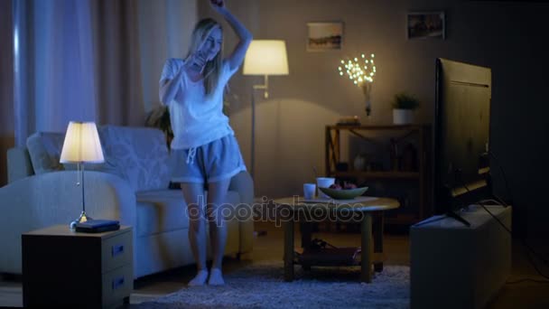 夜美しい女性楽しく踊りながらテレビを見て、また彼女を保持しているスマート フォンの彼女のリビング ルームに. — ストック動画
