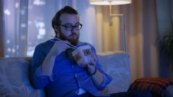 In the Evening Man Sitting on a Couch In His Living Room. He's Eating Asian Food from the Food Container and Watching TV Show. — Stock Video