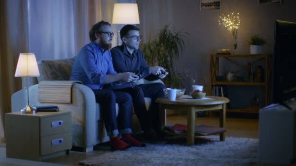 In de avond zitten twee vrienden op een Sofa in de woonkamer en concurrerende videospelletjes spelen. Ze duwen en duwen elkaar vriendelijk. — Stockvideo