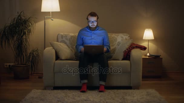 Портрет человека, сидящего на диване в гостиной, ноутбук на коленях. Он работает. Напольные лампы включены . — стоковое видео