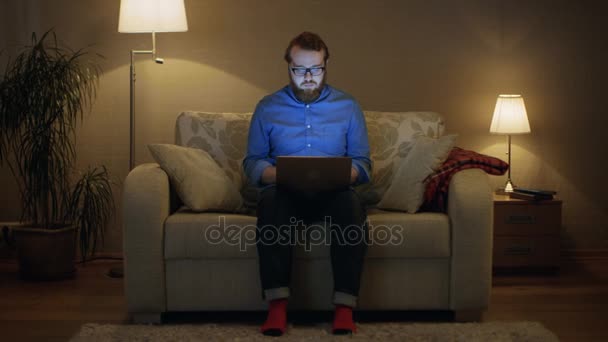 Κατακόρυφος βολή ενός ανθρώπου που κάθεται σε έναν καναπέ στο σαλόνι του Laptop στα γόνατά του. Εργάζεται. Κοιτάζει απευθείας στην κάμερα. Οι λαμπτήρες πατωμάτων είναι ενεργοποιημένο. — Αρχείο Βίντεο