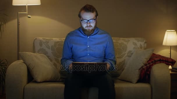 坐在他的客厅里的沙发上，在他的膝盖上的笔记本电脑上的男人的肖像镜头。他的工作。落地灯是转上. — 图库视频影像