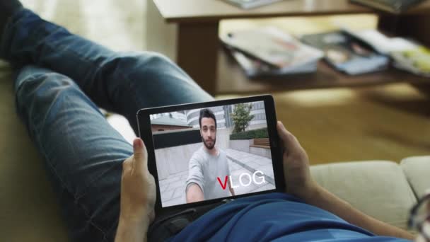 Случайный человек лежит на диване и смотрит модный видеоблог на своем планшетном компьютере. На экране появляется надпись "Vlog" . — стоковое видео