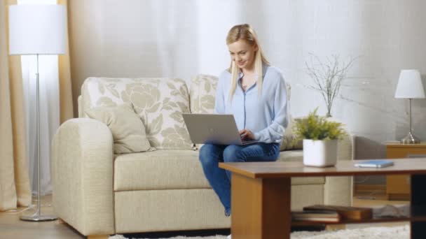 Jeune femme souriante travaillant sur un ordinateur portable. Elle est dans son salon assise sur un canapé et tenant un carnet sur son tour . — Video