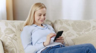 Güzel genç sarışın bir kanepe oturma odasında oturuyor. Gülümseyen, o akıllı telefon kullanır.