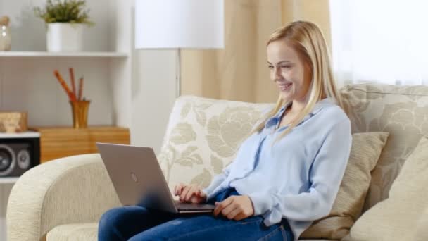 Colpo lungo di una bella donna sorridente seduta su un divano nel suo salotto. Lei lavora sul suo computer portatile che lei tiene sul suo grembo . — Video Stock