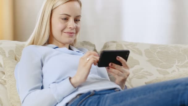 Hermosa joven rubia en su sala de estar se sienta en un sofá, ella ve videos en su teléfono inteligente . — Vídeos de Stock